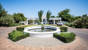 Une fontaine circulaire avec un élément décoratif aquatique au centre, entourée d’une zone pavée et de haies taillées. Au fond, on aperçoit un bâtiment sur lequel est apposée une enseigne indiquant « DE ZALZ ».