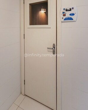 A white door with a small window at the top, a metal handle, and a wall-mounted device with a blue label.
