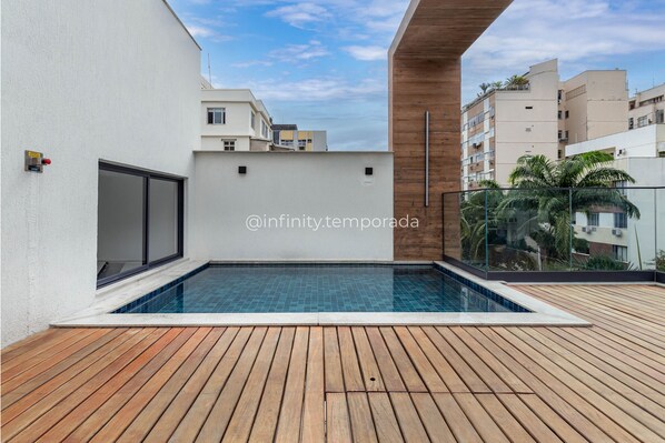 A modern rooftop pool with wooden decking, surrounded by white walls and a clear sky.