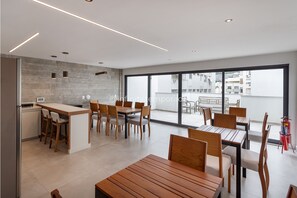 A modern dining area with wooden tables and chairs, a kitchen area with a bar, and large windows offering a view of the city.