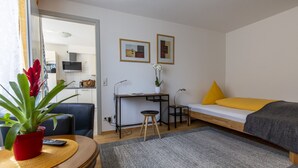 A compact room with a bed, a desk, a chair, a small stool, a potted plant, and a television in the background. - Apartment 'Topas' with Wi-Fi (Freiburg im Breisgau)