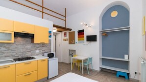 Une cuisine compacte avec des meubles de couleur jaune, un four à micro-ondes et un petit réfrigérateur. On y trouve également un coin repas avec une table blanche et des chaises colorées.