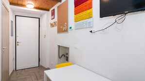 Une petite pièce bien éclairée, avec une porte blanche, un tableau d’affichage en liège où sont accrochées des notes, un téléviseur et une petite table avec une chaise jaune.