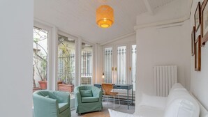 Habitació lluminosa amb parets blanques, un sofà blanc, dues butaques verdes, un faldell trenat i una taula amb cadiretes.