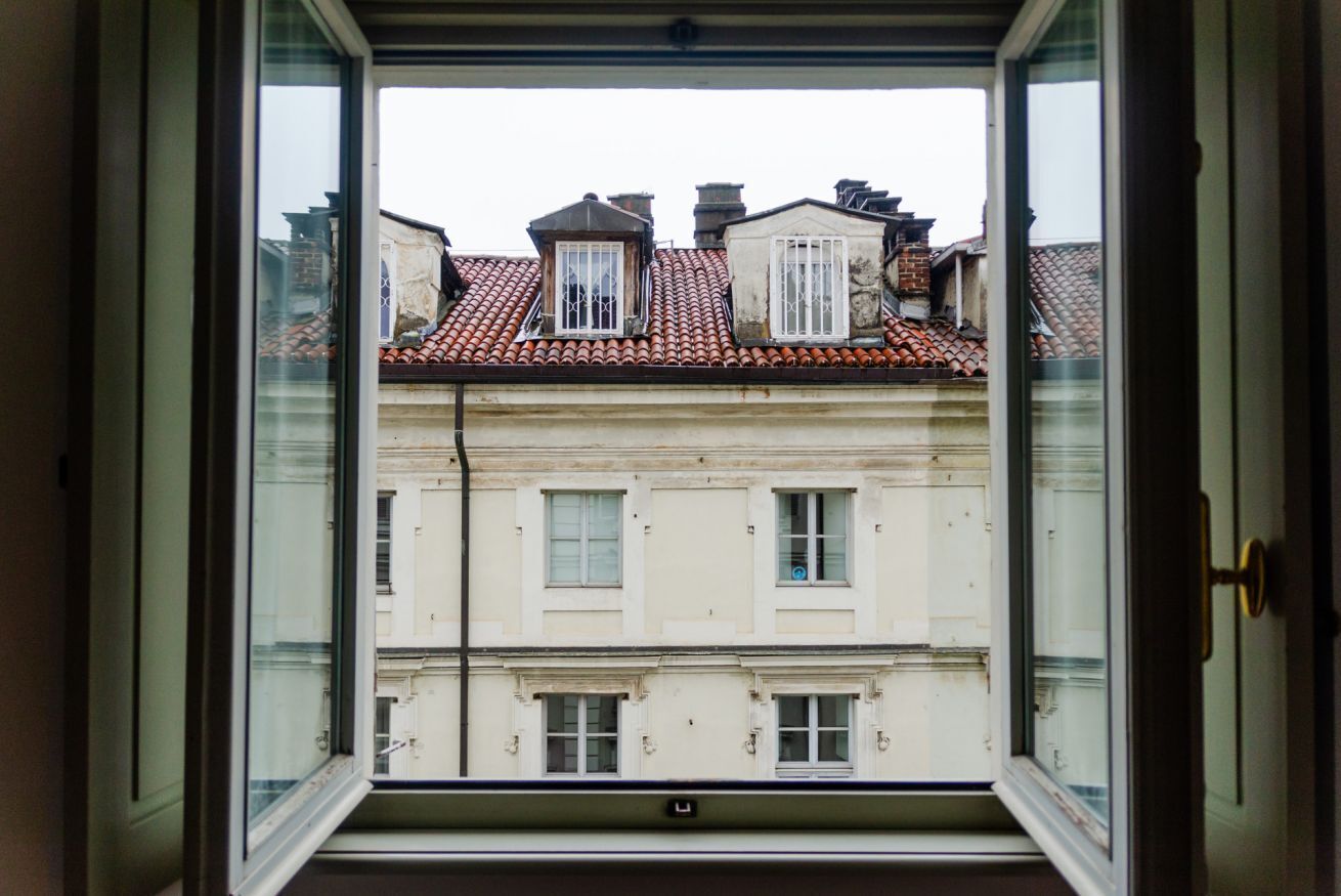Une vue à travers une fenêtre ouverte, donnant sur un immeuble à plusieurs étages, avec un toit recouvert de tuiles rouges et des murs blancs.