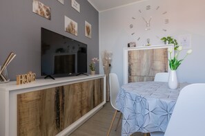 A modern living room with a flat-screen TV, a wooden cabinet, a dining table with a patterned tablecloth, and a wall clock.