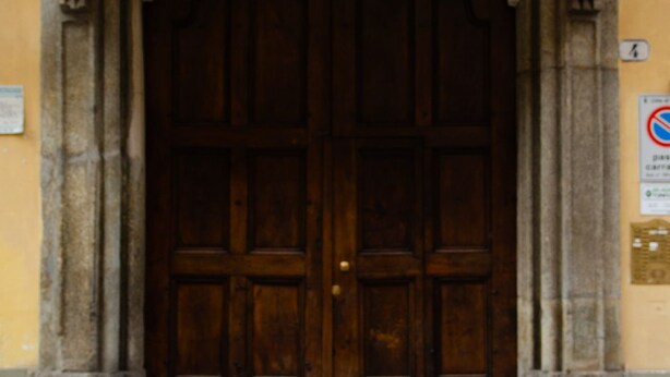 Une grande porte à deux battants en bois, dotée d’un cadre sombre, flanquée de colonnes en pierre, et un panneau indiquant l’interdiction de se garer est fixé au mur, sur la droite.