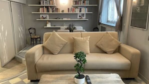 Una sala de estar con un sofá color beige, una pequeña planta en maceta y un control remoto sobre una mesa de centro. Se observan estanterías con libros y objetos decorativos, y una ventana con cortinas.