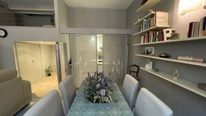 Un comedor con una mesa dispuesta para cuatro personas, una estantería con libros y objetos decorativos, y una puerta corrediza que da a otra habitación.