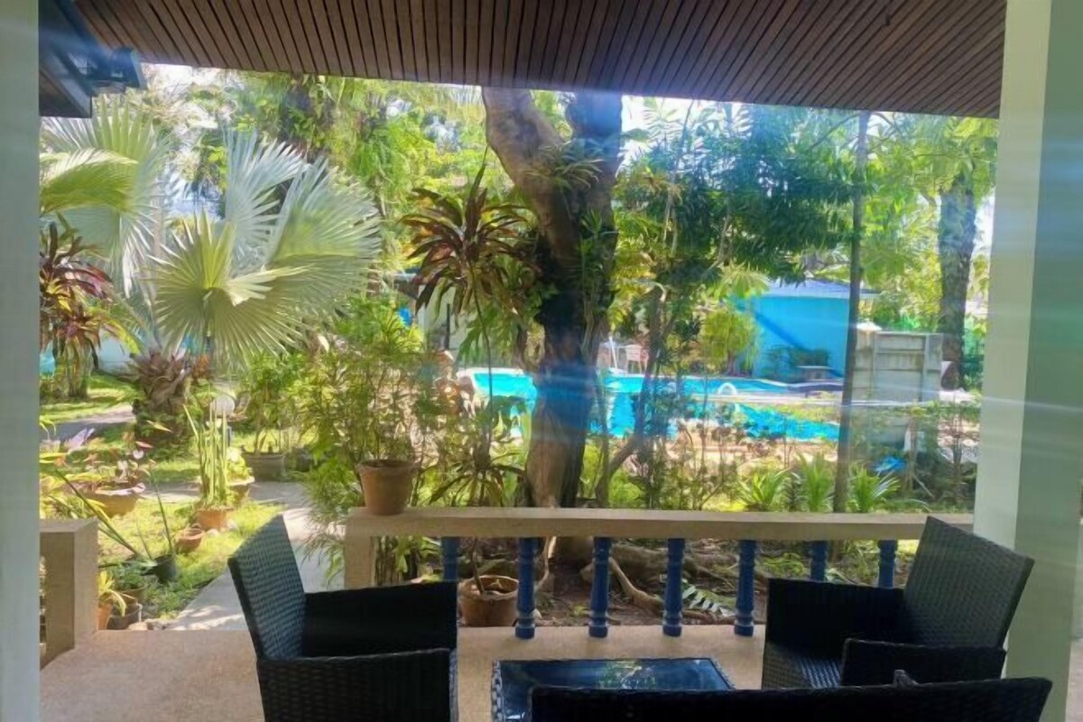 A covered outdoor seating area with wicker furniture, a view of a pool, and lush greenery.