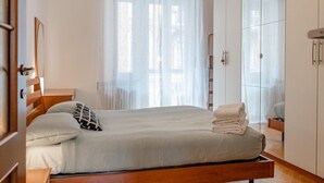 A neatly made bed with a white comforter and pillows, a bedside table with a lamp, and a large window with sheer curtains. - Perrero4 by Happyhostorino (Torino)