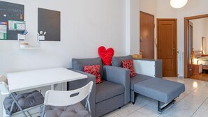 A modern living room with a grey sofa, a white dining table, and a small kitchenette area. - Perrero4 by Happyhostorino (Torino)