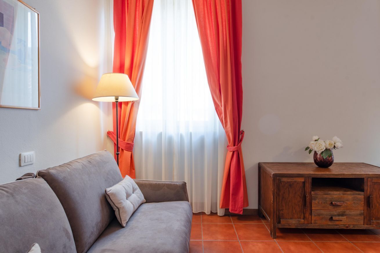 Ein Zimmer mit einem grauen Sofa, einem Holzschrank, einer Lampe und einem Fenster mit roten Vorhängen.