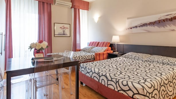 A hotel room with a large bed, a desk with a chair, a nightstand, and a wall-mounted lamp.
