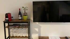 A room with a TV mounted on the wall, a small table with a wine bottle, a potted plant, and a shelf with wine glasses.