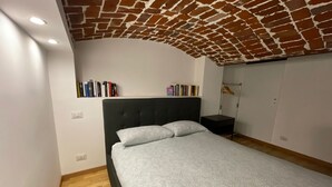 A bedroom with a brick-patterned ceiling, a bed with a grey patterned bedspread, and a bookshelf against the wall.