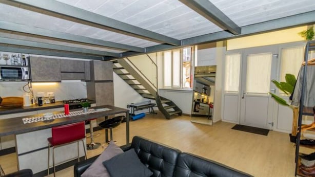 A modern kitchen with a central island, a dining area with a black leather couch, and a staircase leading to an upper level.
