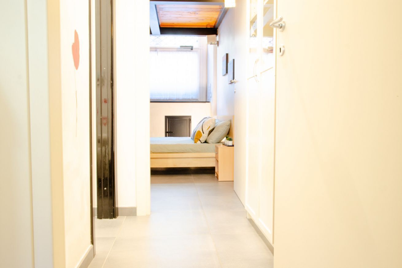 A narrow hallway leading to a bedroom with a bed, a small bedside table, and a window.