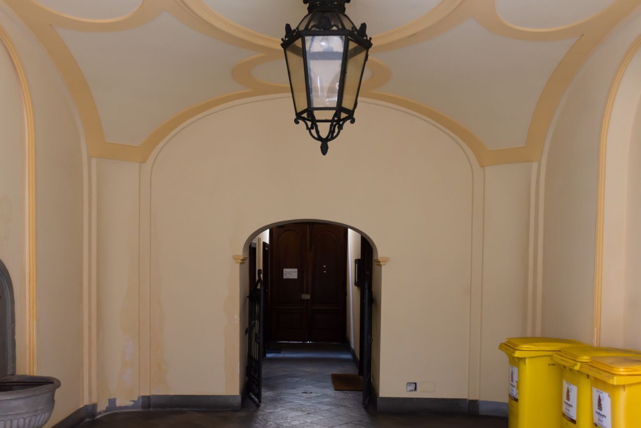 Un pasillo con una puerta de madera al final, un farol negro colgando del techo y un basurero a la derecha.