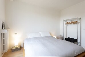 A neatly made bed with white linens, a bedside table with a lamp, a radiator, and a metal rack with hangers and bags. - Attico con vista sulla Consolata (Torino)