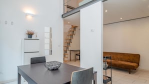 A modern dining area with a dark wooden table, a geometric bowl, and a white cabinet with a plant. A staircase leads to an upper level, and a brown sofa is visible in the background.