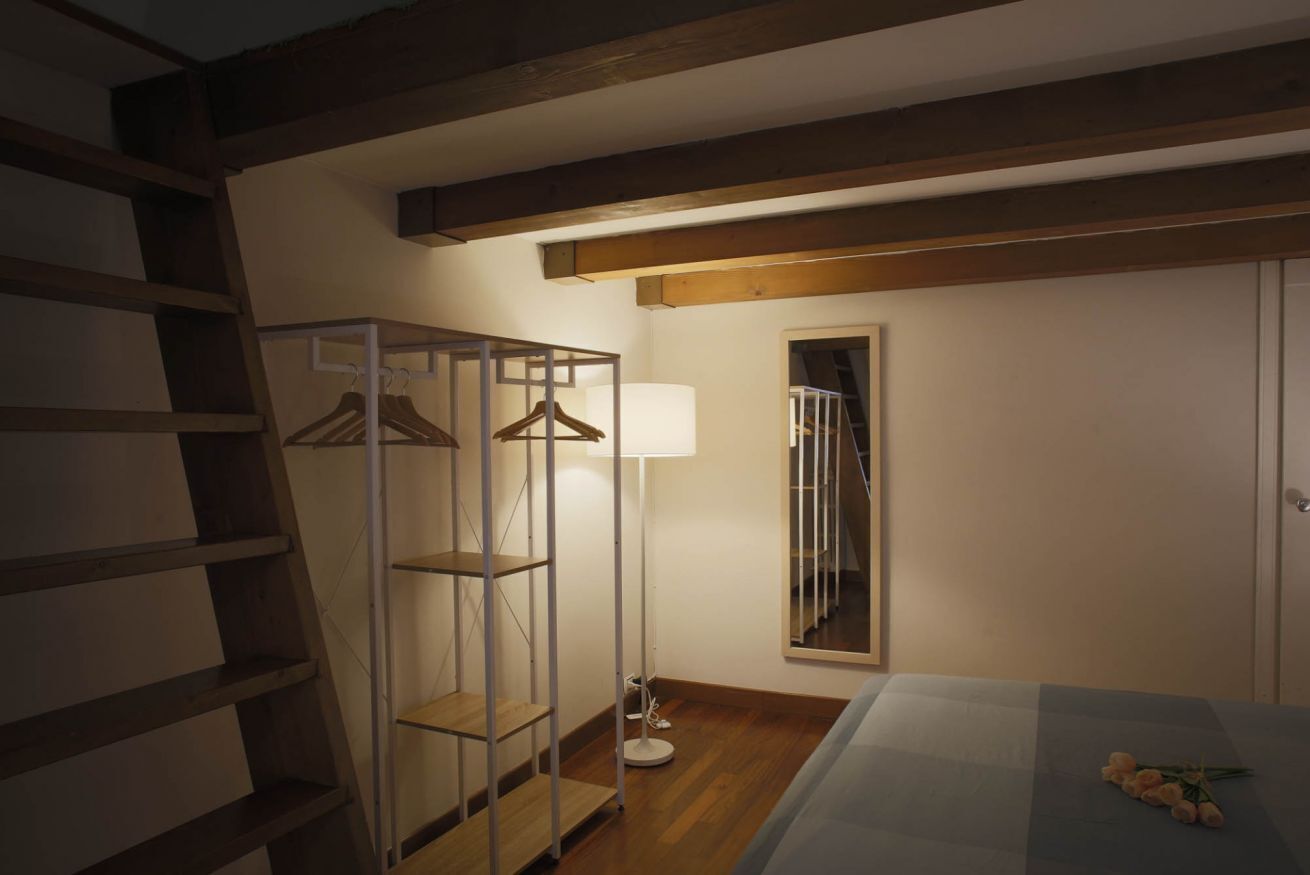 A bedroom with a bed, a wooden ladder, a metal shelving unit, a floor lamp, and a mirror on the wall.