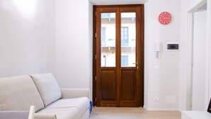 Ein Zimmer mit einer Holztür, einer weißen Wanduhr, einem weißen an der Wand befestigten Telefon und einem beigefarbenen Sofa.