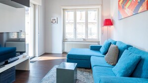 Un salon moderne avec un canapé bleu, un téléviseur à écran plat et une grande fenêtre donnant sur des immeubles.