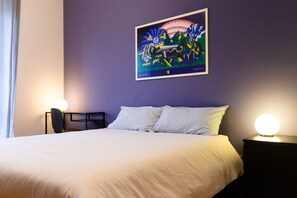 A hotel room with a bed, two bedside lamps, a desk with a chair, and a wall-mounted painting.
