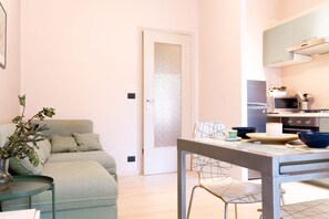 A modern living room with a sofa, a dining table with chairs, a kitchen area with appliances, and a door with a frosted glass panel.
