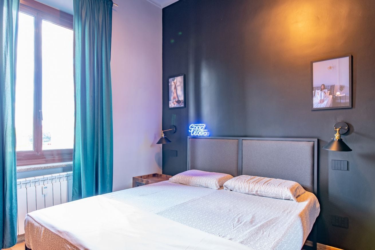 A hotel room with a bed, a window with curtains, a neon sign, and wall-mounted pictures.
