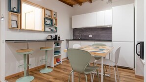 Eine kleine Küche mit weißen Schränken, einer schwarzen Arbeitsplatte und einer Holzbalkendecke. Zwei Hocker und ein Tisch mit Stühlen sind vorhanden.