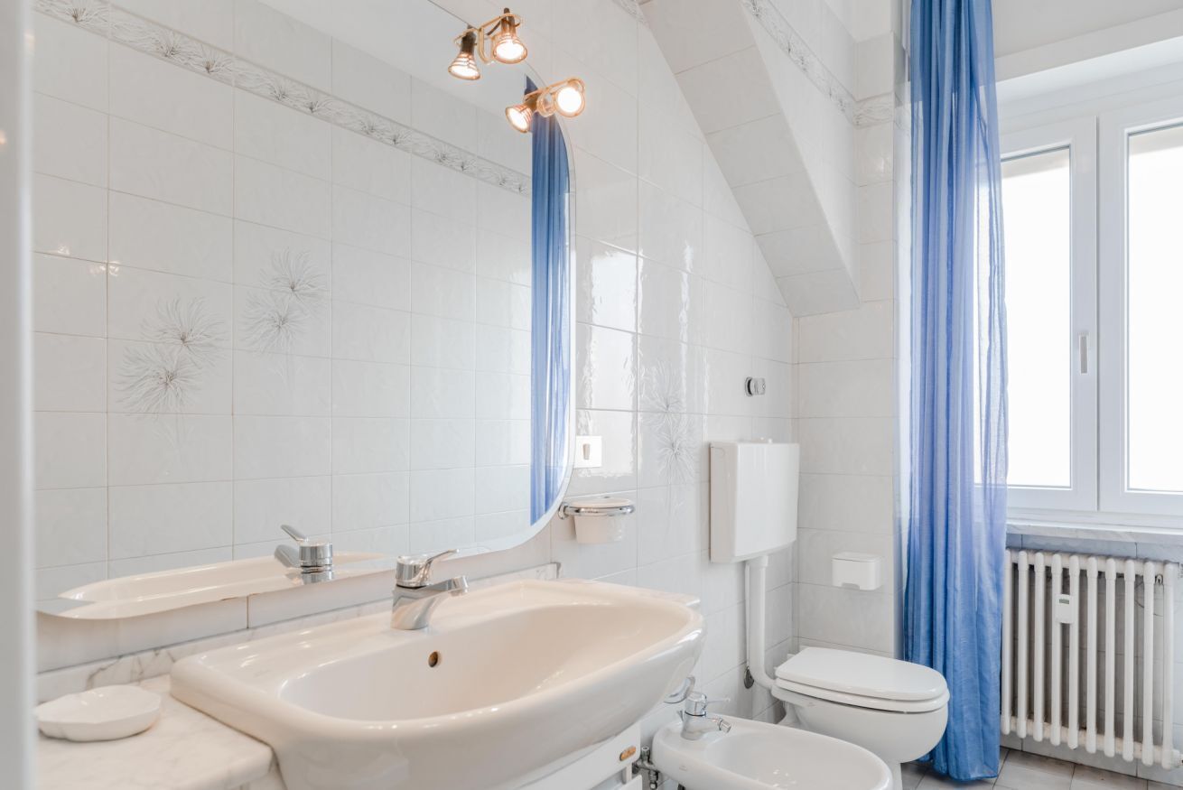 Un bany amb un envàs gran, un vàter i un mirall. Hi ha cortines blaves i un radiador blanc.