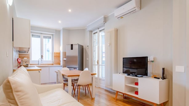 Een moderne woonkamer met een witte bank, een eettafel met stoelen, een televisie op een wit meubelstuk en een keuken in de achtergrond.
