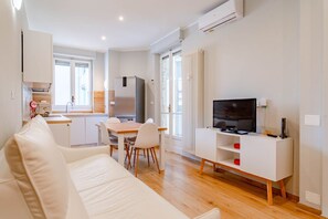 Un saló modern amb un sofà blanc, una taula de dinar amb cadirees, una televisió sobre un moble blanc i una zona de cuina al fons.