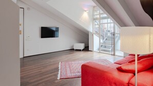 A modern loft bedroom with a sloped ceiling, a flat-screen TV mounted on the wall, a red sofa, and a large window offering a view of the outside. - La Terrazza sui Tetti (Torino)