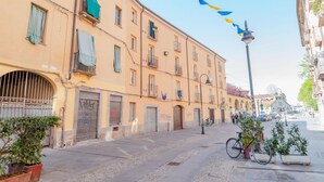A street with a building featuring a large arched entrance, multiple windows, and balconies. There are potted plants and a bicycle parked on the sidewalk. - Casa M&M] Luminosa: Borgo Dora (Torino)