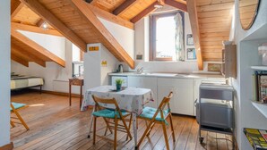 A cozy attic room with wooden beams, a dining table with chairs, a kitchenette, and a window providing natural light. - Casa M&M] Luminosa: Borgo Dora (Torino)