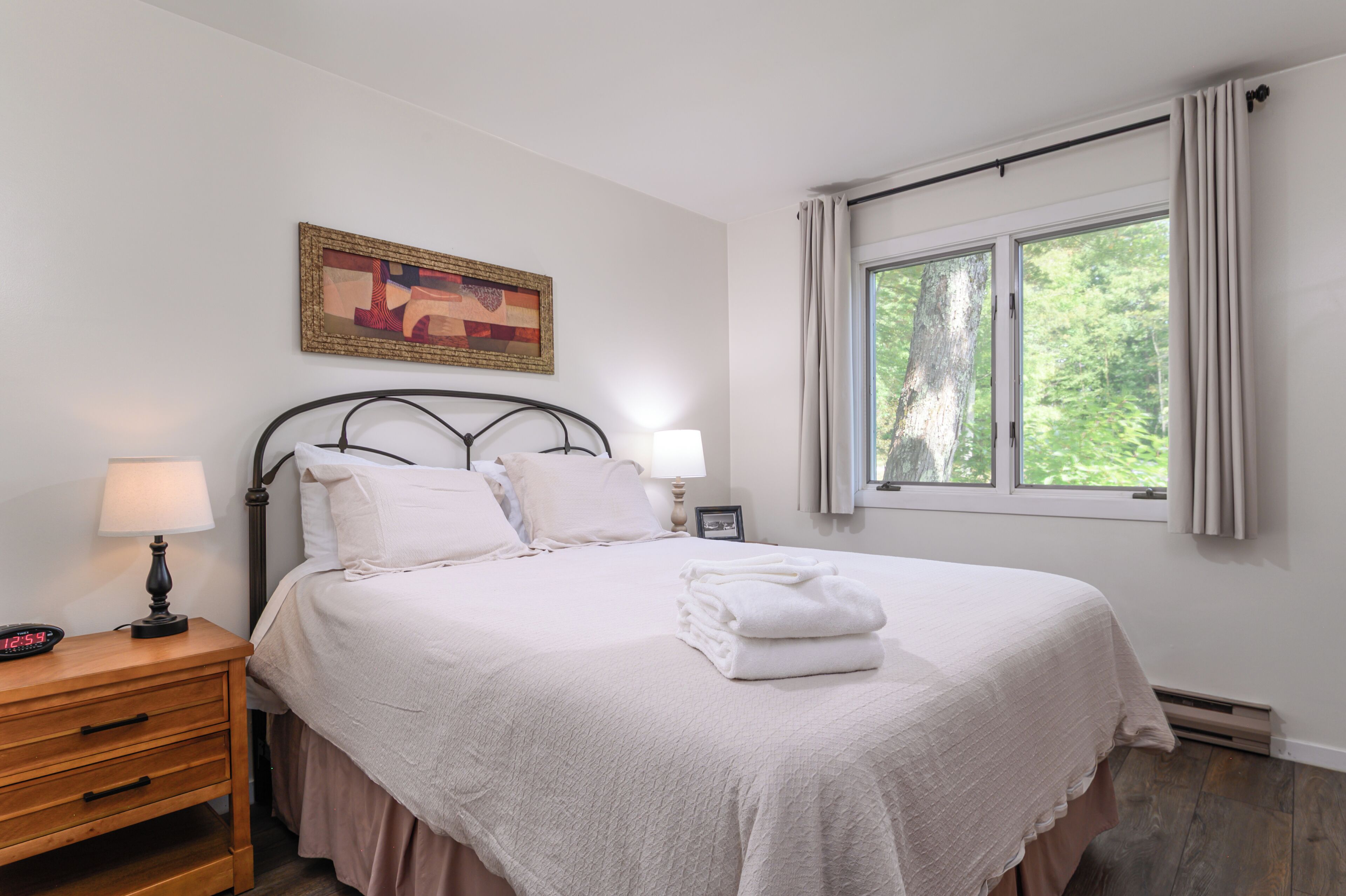 Una cama bien hecha con sábanas blancas y una toalla doblada, una mesita de noche con una lámpara, un cuadro en la pared y una ventana con cortinas.
