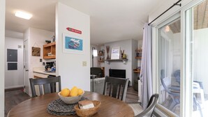 Un espace repas avec une table ronde, des chaises, un saladier de fruits et un panneau indiquant un service de location de matériel de ski.