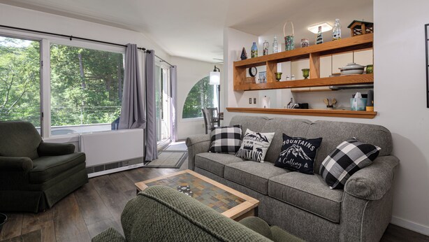 Un salon chaleureux avec un canapé gris, un fauteuil vert et une table basse en bois. Une fenêtre avec des rideaux laisse entrer la lumière naturelle, et une étagère au-dessus du canapé expose des objets décoratifs.