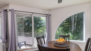 Un coin repas avec une table ronde, des chaises, un saladier rempli de fruits et une vue dégagée sur l’extérieur grâce à une grande fenêtre.