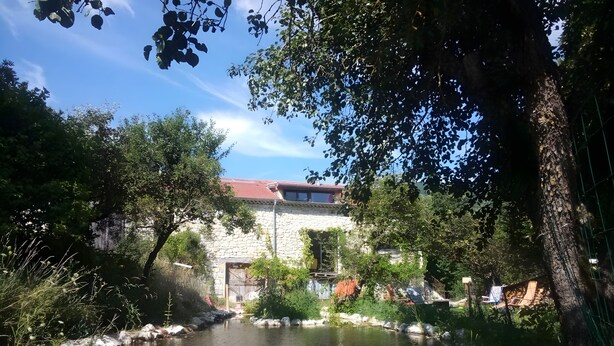 A stone building with a red roof, surrounded by greenery and a calm water body. - Ferme des Montagnes Bleues – Guest Room with Breakfast in the Peaceful Drôme Mountains (Bellegarde-en-Diois)