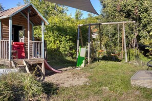Una casita de madera con un tobogán, rodeada de árboles y una zona de césped.