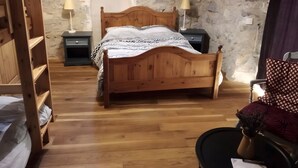 A wooden bed with a patterned bedspread, two bedside tables with lamps, a stone wall, and a window with a view of the outside. - Ferme des Montagnes Bleues – Guest Room with Breakfast in the Peaceful Drôme Mountains (Bellegarde-en-Diois)
