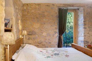 Una habitación con paredes de piedra, una cama de madera con colcha estampada, una lámpara de pie y una ventana con cortina.