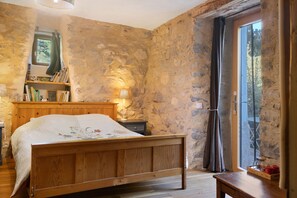 Un dormitorio con una cama de madera, una mesita de noche, una lámpara, una estantería y vistas a través de una ventana con cortinas.