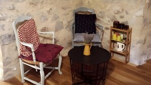 A rustic stone wall, a white wooden chair with a red cushion, a blue cushioned chair, a small round table with a yellow vase, and a wooden shelf with kitchen items. - Ferme des Montagnes Bleues – Guest Room with Breakfast in the Peaceful Drôme Mountains (Bellegarde-en-Diois)