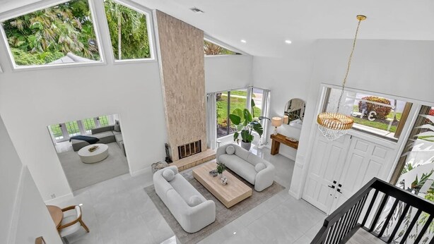 A spacious living area with a large sofa set, a coffee table, and a fireplace. There are large windows offering views of greenery outside. - Gorgeous Modern Beach Heated Pool House Sleeps 8 Central (Delray Beach)