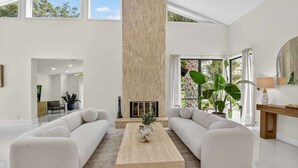 A spacious living room with a high ceiling, a fireplace, and large windows offering a view of greenery. - Gorgeous Modern Beach Heated Pool House Sleeps 8 Central (Delray Beach)